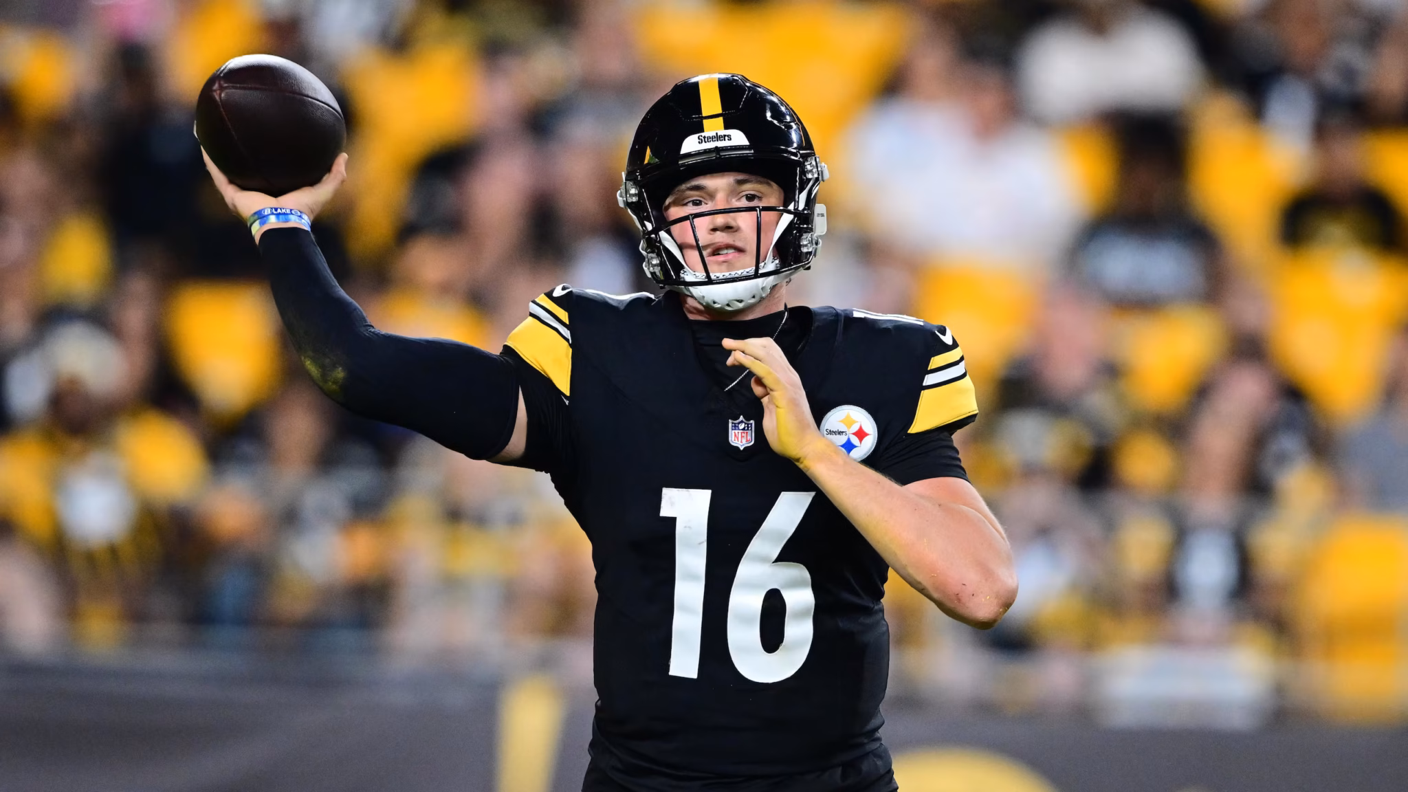 Quarterback in a Steelers uniform winds up to throw a football with a crowded stadium in yellow seats behind him.